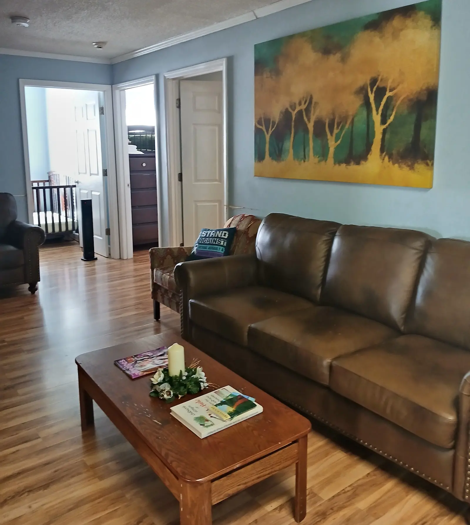 Shelter common area Living room area with a brown leather sofa, wooden coffee table, and large painting of trees on the wall. Two doorways lead to other rooms.