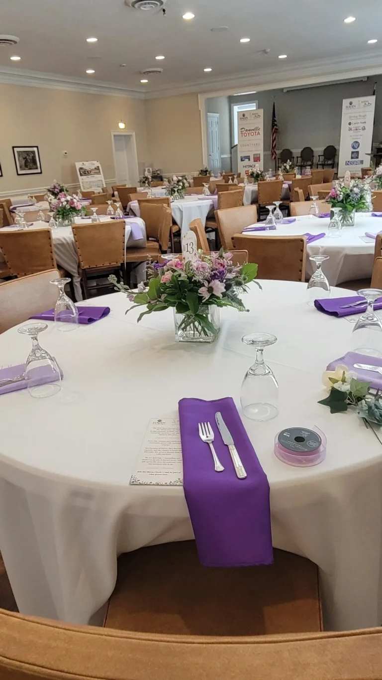 May show: Round banquet tables with white tablecloths, purple napkins, floral centerpieces, and place settings are arranged in a decorated event hall.